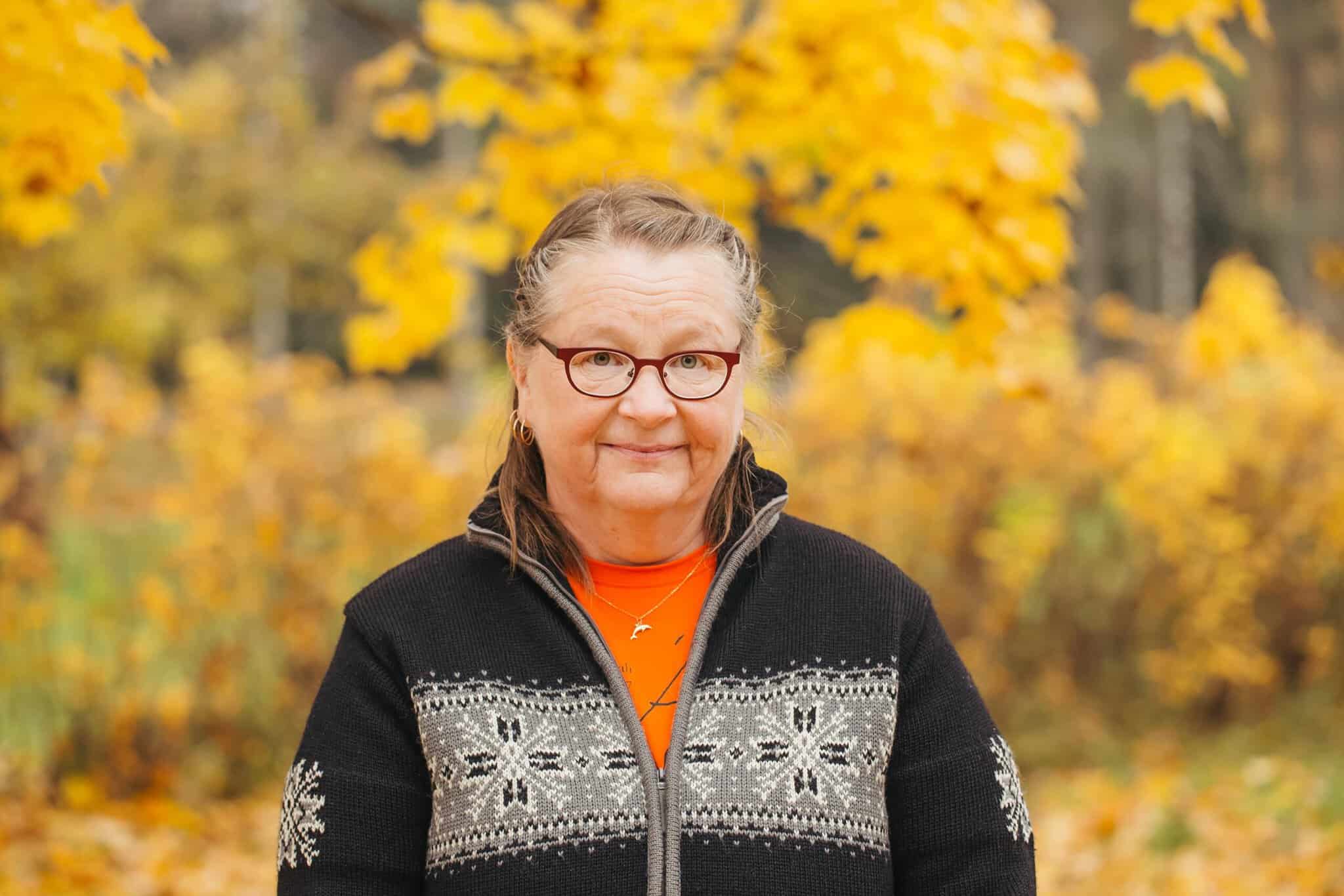 Hymyilevä nainen, jolla silmälasti ja harmaa neule, ruskalehtiä taustalla.